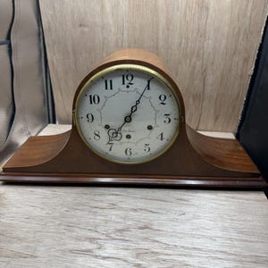 May include: A wooden mantel clock with a curved top and a wide base. The clock face is white with black Arabic numerals, a gold-toned metal bezel, and black ornate hands. The text Seth Thomas is printed in black script on the center of the dial.