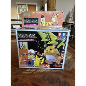 Könnte beinhalten: Eine silberne rechteckige Box mit einem Cartoon-Charakter Pikachu und Blitzakzenten. Die Box hat japanischen Text und das TOMY-Logo. Die Box hat ein gelbes und schwarzes Design. Die Box steht auf einer Holzoberfläche.