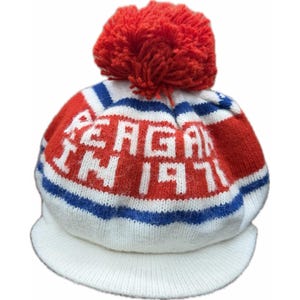 K&ouml;nnte beinhalten: Eine Strickm&uuml;tze mit Schirm, verziert mit einem roten Bommel. Die M&uuml;tze ist wei&szlig;, rot und blau, mit dem Text "REAGAN IN 1971" in wei&szlig;en Buchstaben. Der Schirm ist wei&szlig;, f&uuml;r einen klassischen Look.