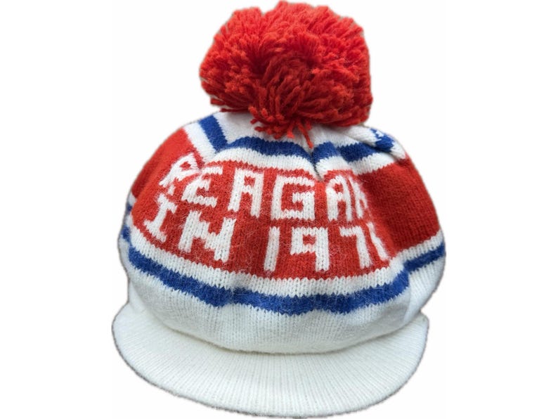 K&ouml;nnte beinhalten: Eine Strickm&uuml;tze mit Schirm, verziert mit einem roten Bommel. Die M&uuml;tze ist wei&szlig;, rot und blau, mit dem Text "REAGAN IN 1971" in wei&szlig;en Buchstaben. Der Schirm ist wei&szlig;, f&uuml;r einen klassischen Look.