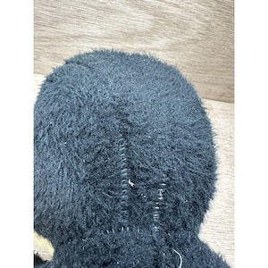 May include: Close-up of a black, fuzzy plush toy. The toy has a stitched seam running down the center. The fur is dense and covers the entire surface, giving it a soft appearance. The background is a light brown wooden surface.