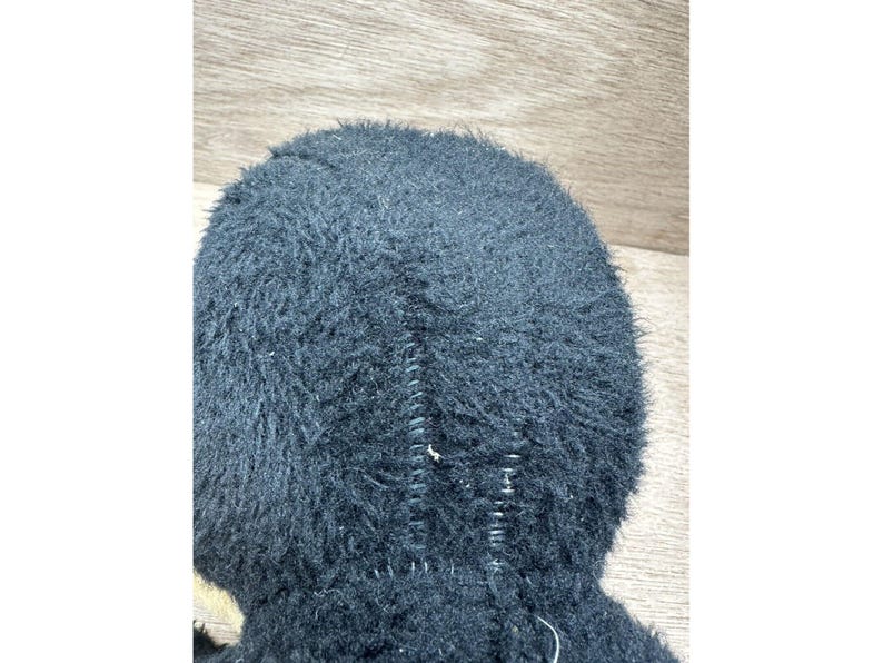 May include: Close-up of a black, fuzzy plush toy. The toy has a stitched seam running down the center. The fur is dense and covers the entire surface, giving it a soft appearance. The background is a light brown wooden surface.