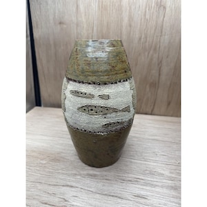 May include: A handcrafted ceramic vase with a textured surface and a unique fish design. The vase features a brown and beige color scheme, with the fish design in a lighter beige color. The vase is approximately 10 inches tall.