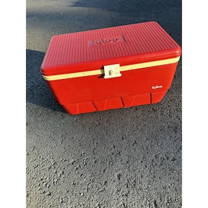 May include: A red Igloo cooler with a white latch and a cream-colored trim. The top of the cooler has a textured surface with the Igloo logo embossed. The cooler is sitting on a gray asphalt surface.