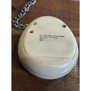 May include: Cream-colored, egg-shaped electronic toy with a silver chain. The toy has a small screen and buttons. Text on the back reads "(C) 1997 MGA ENTERTAINMENT All Rights Reserved".