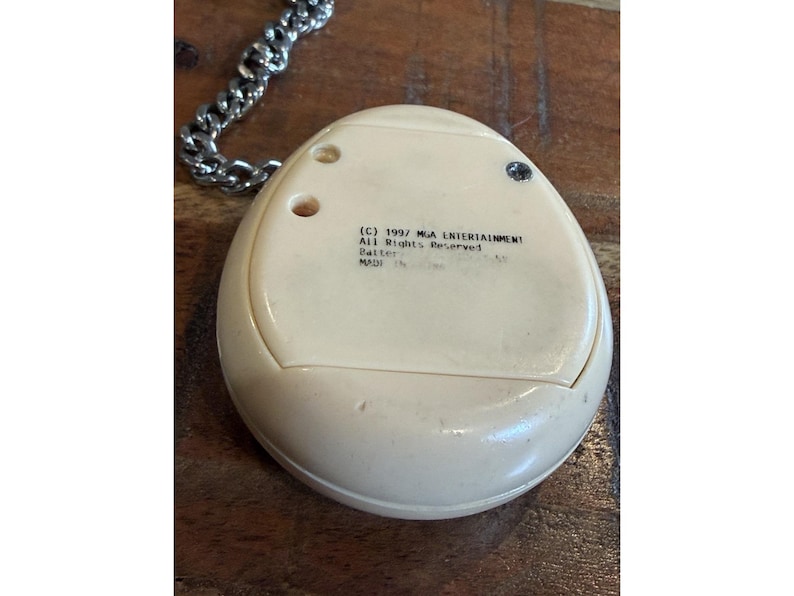 May include: Cream-colored, egg-shaped electronic toy with a silver chain. The toy has a small screen and buttons. Text on the back reads "(C) 1997 MGA ENTERTAINMENT All Rights Reserved".