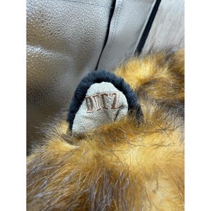 May include: Close-up of a plush toy ear with brown and tan fur. The ear has a black trim and a beige inner ear with the embroidered letters "DITZ". The toy appears to be a stuffed animal.