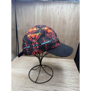May include: A five-panel baseball cap featuring a black base with a vibrant geometric pattern in shades of red, orange, and teal. The cap has a solid black brim and a black adjustable strap at the back, displayed on a black wire stand.