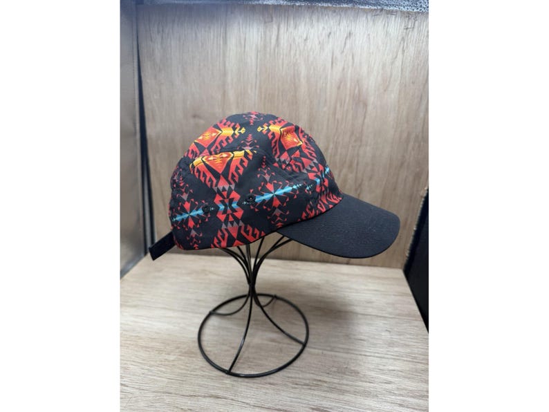 May include: A five-panel baseball cap featuring a black base with a vibrant geometric pattern in shades of red, orange, and teal. The cap has a solid black brim and a black adjustable strap at the back, displayed on a black wire stand.