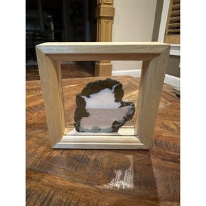 May include: A natural stone specimen encased in a light-colored wooden frame. The stone features a mix of brown, white, and pink hues. The frame has a simple, rectangular design, creating a modern display piece.