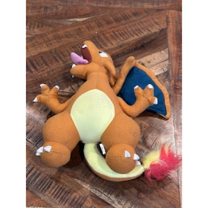 May include: A plush Charizard toy in shades of orange, yellow, and blue. The toy has a light yellow belly, blue wings, and a red and yellow tail. The mouth is open, revealing a pink tongue.