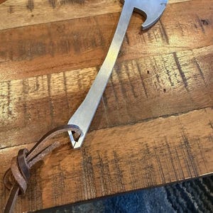 May include: A silver-colored metal bottle opener shaped like a small axe. The handle features a hole at the base with a brown leather cord threaded through it, tied in a knot. The metal surface has a brushed finish and a distinct axe head design.