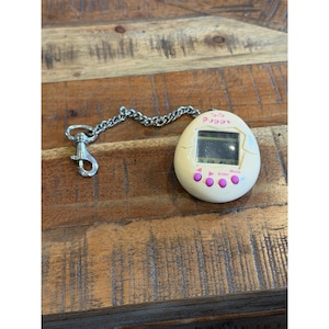 May include: A vintage Tamagotchi digital pet toy in an egg shape, cream-colored with a small screen and pink buttons. It has a silver chain and clasp for attachment. The screen displays a digital interface.