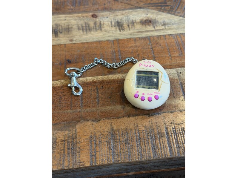 May include: A vintage Tamagotchi digital pet toy in an egg shape, cream-colored with a small screen and pink buttons. It has a silver chain and clasp for attachment. The screen displays a digital interface.
