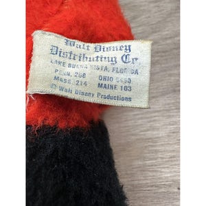 May include: Close-up of a vintage plush toy with a red top and black bottom. A tag reads "Walt Disney Distributing Co." with locations including Lake Buena Vista, Florida, and Ohio. The tag also includes "Walt Disney Productions."
