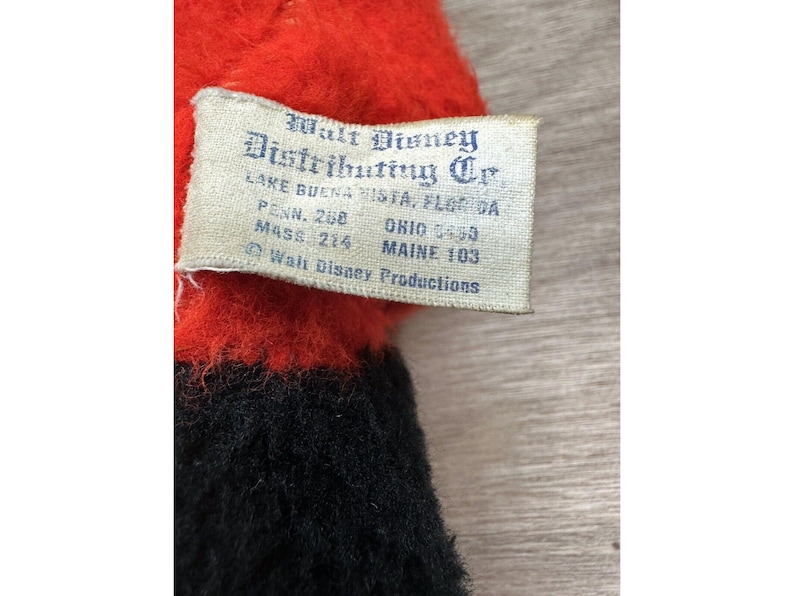 May include: Close-up of a vintage plush toy with a red top and black bottom. A tag reads "Walt Disney Distributing Co." with locations including Lake Buena Vista, Florida, and Ohio. The tag also includes "Walt Disney Productions."