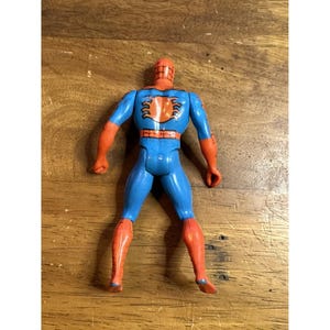 Puede incluir: Una figura de acci&oacute;n de Spider-Man vista desde atr&aacute;s, con cuerpo azul, extremidades naranjas y un prominente emblema de ara&ntilde;a naranja en el centro de la espalda. La figura tiene una cabeza roja con un patr&oacute;n de telara&ntilde;a negro y un cintur&oacute;n rojo.