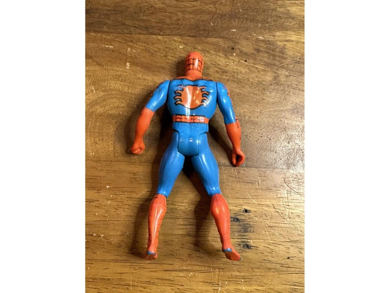 Puede incluir: Una figura de acci&oacute;n de Spider-Man vista desde atr&aacute;s, con cuerpo azul, extremidades naranjas y un prominente emblema de ara&ntilde;a naranja en el centro de la espalda. La figura tiene una cabeza roja con un patr&oacute;n de telara&ntilde;a negro y un cintur&oacute;n rojo.