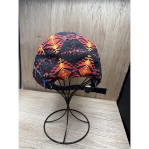 May include: A black baseball cap featuring a repeating geometric pattern in shades of red, orange, and yellow. The cap includes a black fabric strap at the back with a metallic buckle for adjustment, displayed on a thin black wire stand.