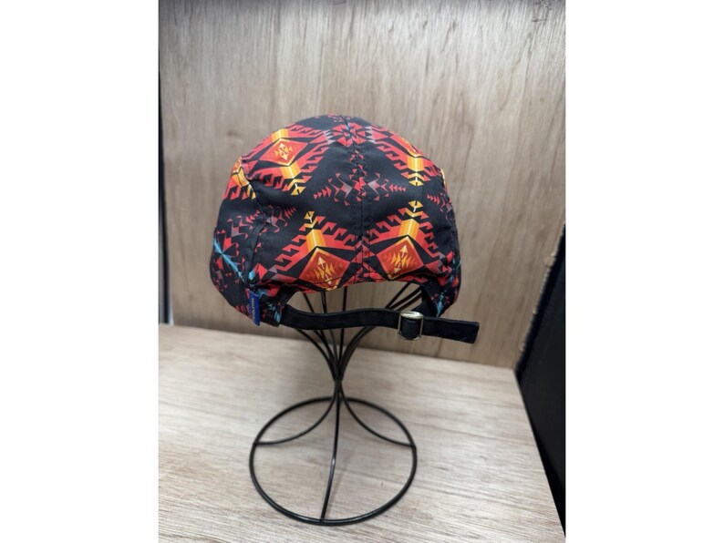 May include: A black baseball cap featuring a repeating geometric pattern in shades of red, orange, and yellow. The cap includes a black fabric strap at the back with a metallic buckle for adjustment, displayed on a thin black wire stand.