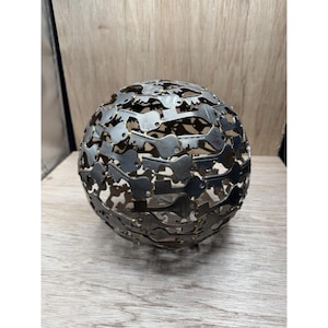 Puede incluir: Una esfera decorativa de metal compuesta por numerosos recortes en forma de llave. La esfera tiene un acabado metálico oscuro y se asienta sobre una superficie de madera. El diseño crea una estructura abierta y aireada.