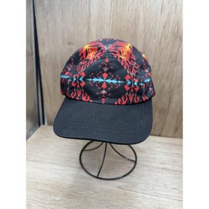 May include: A five-panel cap featuring a black base with a vibrant geometric pattern in shades of red, orange, and light blue. The hat has a solid black flat brim and a structured crown, showcasing a repeating symmetrical design across the fabric.