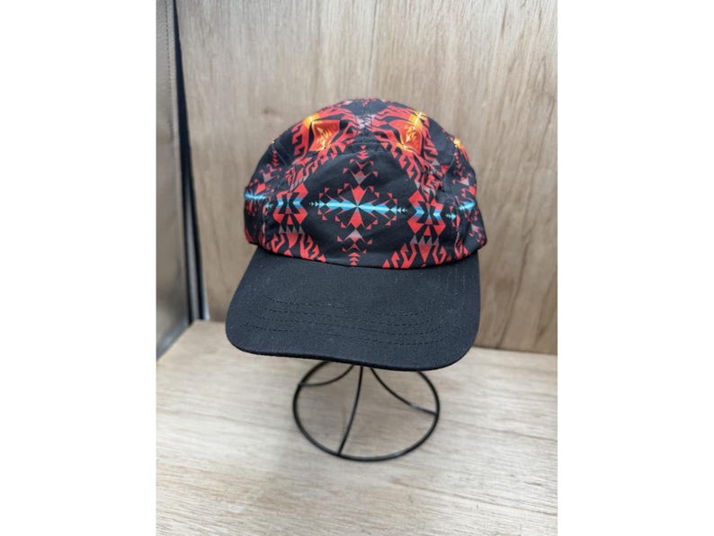 May include: A five-panel cap featuring a black base with a vibrant geometric pattern in shades of red, orange, and light blue. The hat has a solid black flat brim and a structured crown, showcasing a repeating symmetrical design across the fabric.