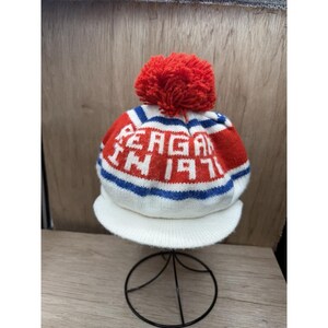 K&ouml;nnte beinhalten: Eine gestrickte M&uuml;tze in Wei&szlig;, Rot und Blau mit dem Text "REAGAN IN 1971" in Wei&szlig;. Sie hat einen roten Bommel und einen wei&szlig;en Schirm. Die M&uuml;tze wird auf einem schwarzen Metallst&auml;nder pr&auml;sentiert.