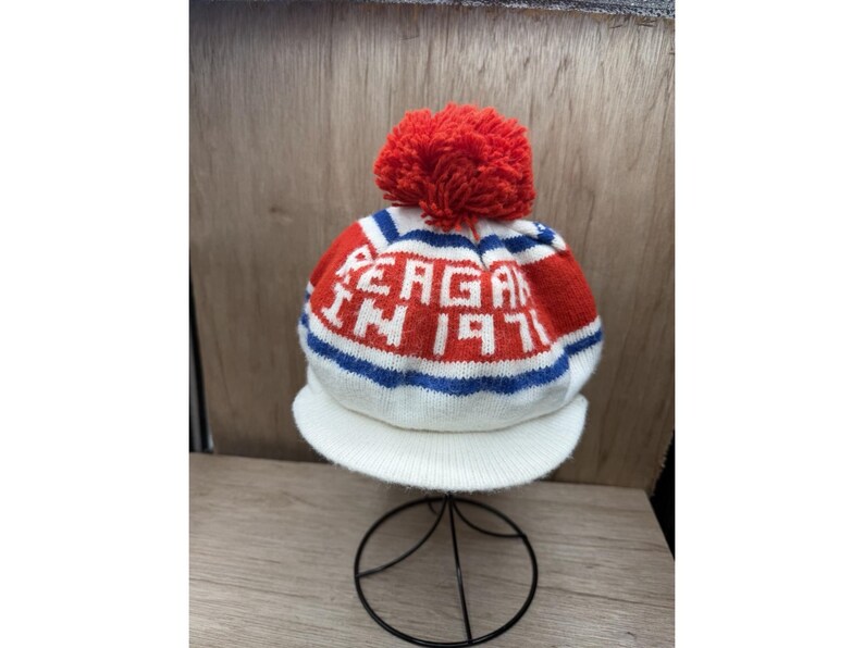 K&ouml;nnte beinhalten: Eine gestrickte M&uuml;tze in Wei&szlig;, Rot und Blau mit dem Text "REAGAN IN 1971" in Wei&szlig;. Sie hat einen roten Bommel und einen wei&szlig;en Schirm. Die M&uuml;tze wird auf einem schwarzen Metallst&auml;nder pr&auml;sentiert.