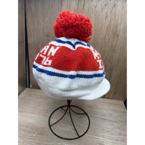 K&ouml;nnte beinhalten: Eine wei&szlig;e Strickm&uuml;tze mit einem roten Bommel. Die M&uuml;tze hat ein rot-wei&szlig;es Design mit dem Text "AN 76" in Wei&szlig;. Blaue Streifen akzentuieren das Design. Die M&uuml;tze hat einen kleinen Schirm.