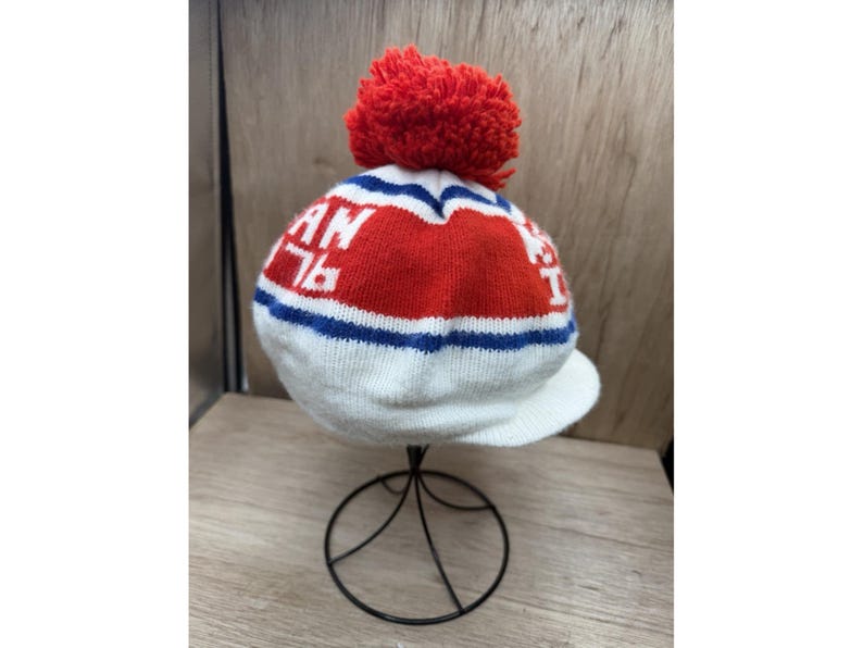 K&ouml;nnte beinhalten: Eine wei&szlig;e Strickm&uuml;tze mit einem roten Bommel. Die M&uuml;tze hat ein rot-wei&szlig;es Design mit dem Text "AN 76" in Wei&szlig;. Blaue Streifen akzentuieren das Design. Die M&uuml;tze hat einen kleinen Schirm.