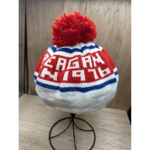 K&ouml;nnte beinhalten: Eine gestrickte M&uuml;tze mit einem roten Bommel. Die M&uuml;tze tr&auml;gt den Namen "REAGAN" und die Nummer "76" in wei&szlig;en Buchstaben auf rotem Grund, mit blauen und wei&szlig;en Streifen. Die M&uuml;tze wird auf einem schwarzen Metallst&auml;nder pr&auml;sentiert.