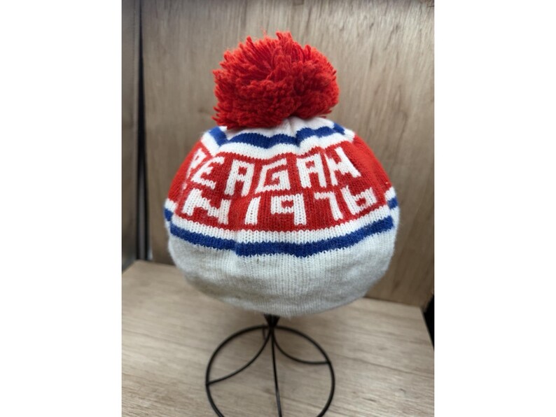 K&ouml;nnte beinhalten: Eine gestrickte M&uuml;tze mit einem roten Bommel. Die M&uuml;tze tr&auml;gt den Namen "REAGAN" und die Nummer "76" in wei&szlig;en Buchstaben auf rotem Grund, mit blauen und wei&szlig;en Streifen. Die M&uuml;tze wird auf einem schwarzen Metallst&auml;nder pr&auml;sentiert.