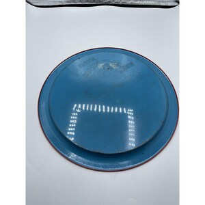 May include: A round, blue metal tray with a red rim. The tray has a slightly worn appearance, with some visible scratches and imperfections. The design is simple and functional, suitable for serving or display.