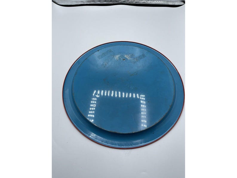 May include: A round, blue metal tray with a red rim. The tray has a slightly worn appearance, with some visible scratches and imperfections. The design is simple and functional, suitable for serving or display.