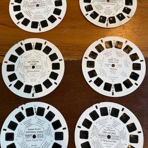 Może przedstawiać: Sześć okrągłych białych krążków View-Master z czarnymi prostokątnymi ramkami na zdjęcia i nadrukowanym tekstem. Krążki przedstawiają różne tematy, w tym Scenic U.S.A., Prehistoric Animals, New Orleans Square, Grand Teton National Park, United States National Parks oraz Walt Disney's Chip and Dale in Flying Saucer Pilots.