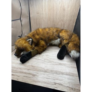 May include: A soft toy fox in shades of brown, orange, and white, lying on a wooden surface. The toy has black paws and a fluffy tail. The fox is in a relaxed pose, with its eyes closed.