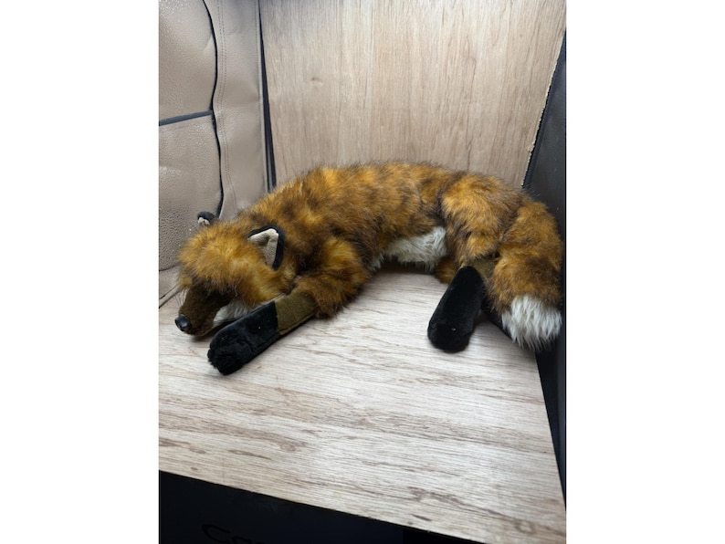 May include: A soft toy fox in shades of brown, orange, and white, lying on a wooden surface. The toy has black paws and a fluffy tail. The fox is in a relaxed pose, with its eyes closed.