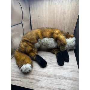 May include: A plush fox toy with brown and orange fur, white chest and tail tip, and black paws. The stuffed animal is lying down on a wooden surface.