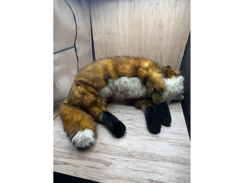 May include: A plush fox toy with brown and orange fur, white chest and tail tip, and black paws. The stuffed animal is lying down on a wooden surface.