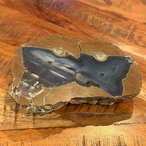 May include: A polished, irregularly shaped stone with a dark gray and white center, framed by a band of tan and brown. The stone's surface has natural patterns and textures, suggesting it is a decorative item or a unique piece of art.