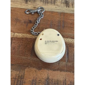 May include: A vintage off-white handheld electronic game with a silver chain and clasp. The game has a rounded, egg-like shape with small buttons and text on the back. The chain is attached to the top of the game.
