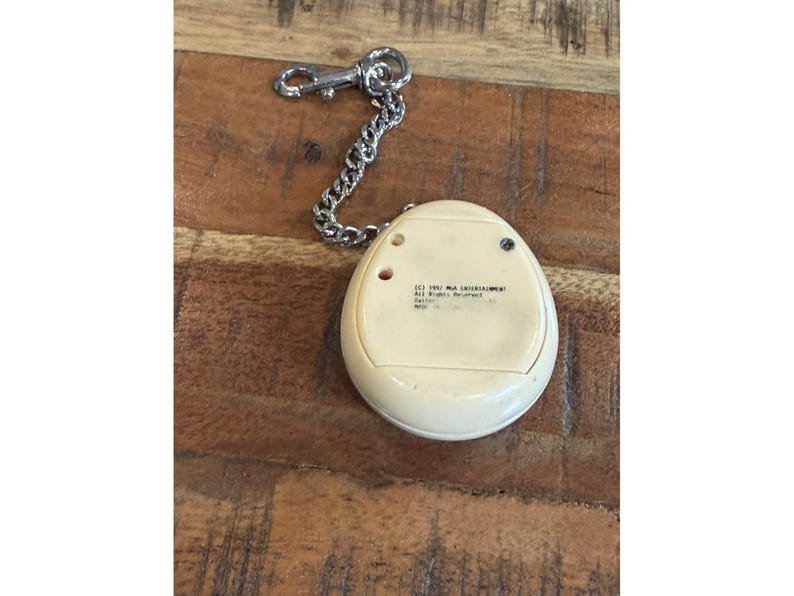 May include: A vintage off-white handheld electronic game with a silver chain and clasp. The game has a rounded, egg-like shape with small buttons and text on the back. The chain is attached to the top of the game.