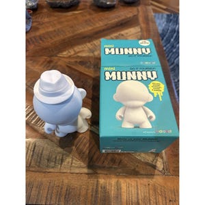 May include: A light blue Munny figure wearing a white hat and a white cape, next to a teal box with the words "mini MUNNY DO IT YOURSELF" printed on it. The box features a white Munny figure and yellow accents.