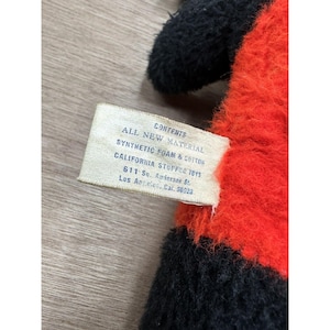 May include: Close-up of a stuffed toy with black and red fur. A tag reads "CONTENTS ALL NEW MATERIAL SYNTHETIC FOAM & COTTON CALIFORNIA STUFFED TOYS 611 So. Andersen St. Los Angeles, Cal. 90023."