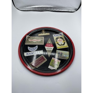 May include: A vintage black serving tray with a red rim, featuring various liquor brand labels. Labels include "Martell", "Drambuie", "Gilbey's Gin", and "Martini Vermouth". The tray has a gold emblem at the top.