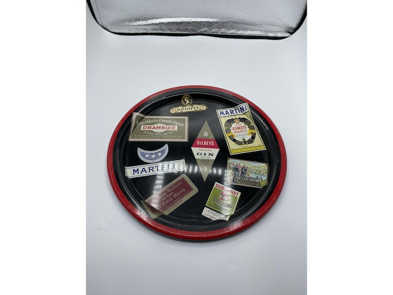 May include: A vintage black serving tray with a red rim, featuring various liquor brand labels. Labels include "Martell", "Drambuie", "Gilbey's Gin", and "Martini Vermouth". The tray has a gold emblem at the top.