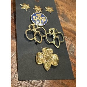 May include: A collection of vintage Girl Scout pins and badges on a black card. The pins include gold star-shaped pins, a blue and gold emblem, two gold figure pins, and a gold trefoil pin with an eagle.