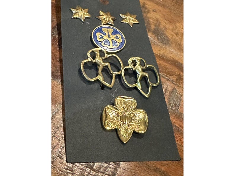 May include: A collection of vintage Girl Scout pins and badges on a black card. The pins include gold star-shaped pins, a blue and gold emblem, two gold figure pins, and a gold trefoil pin with an eagle.