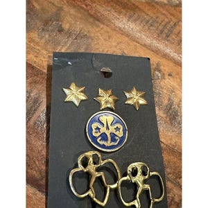 May include: A black card displays Girl Scout pins and badges. Three gold star pins are at the top. Below are a blue and gold round badge and two gold Girl Scout silhouette pins. The items are on a wooden surface.